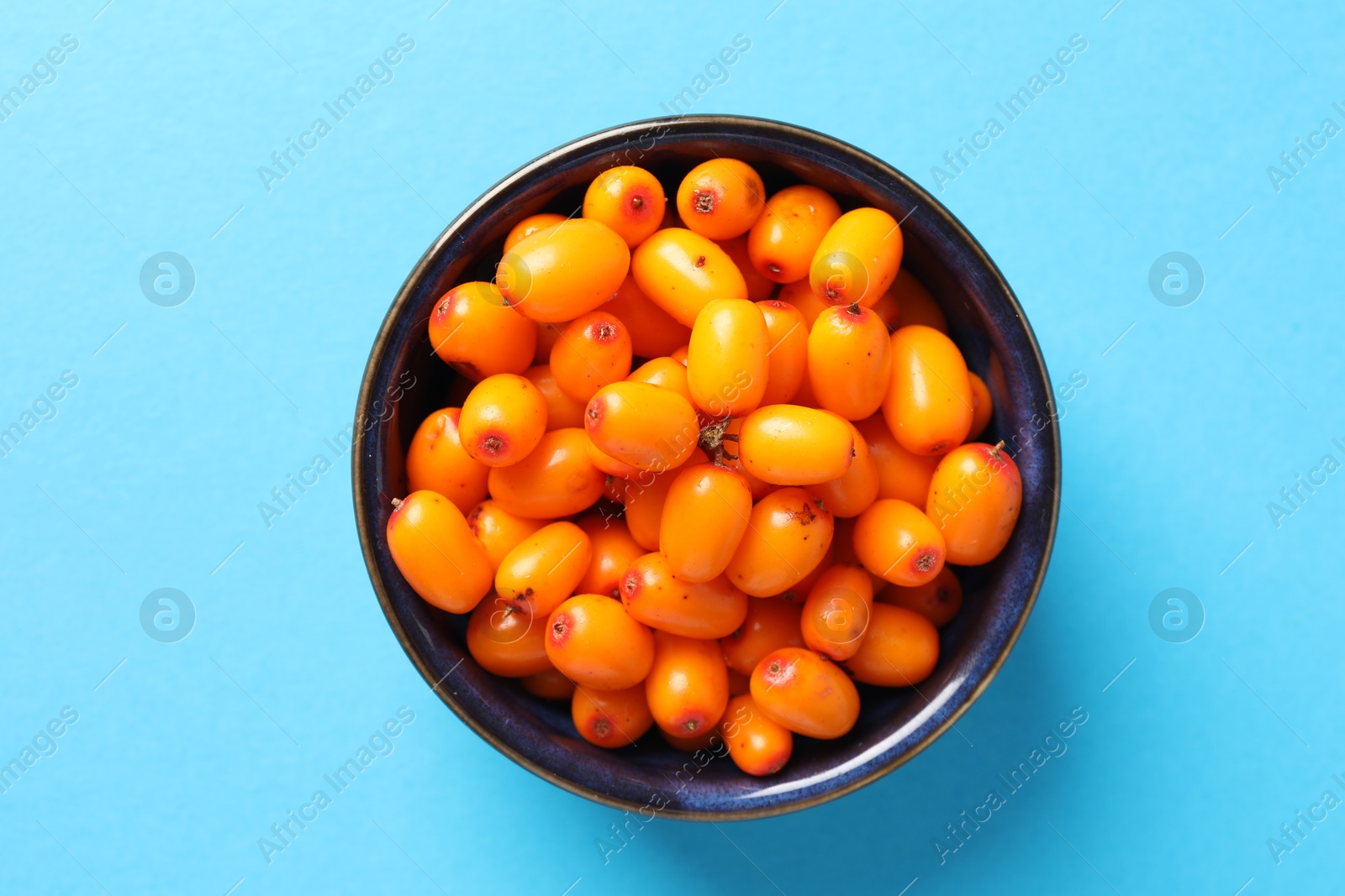 Photo of Ripe sea buckthorn berries in bowl on light blue background, top view