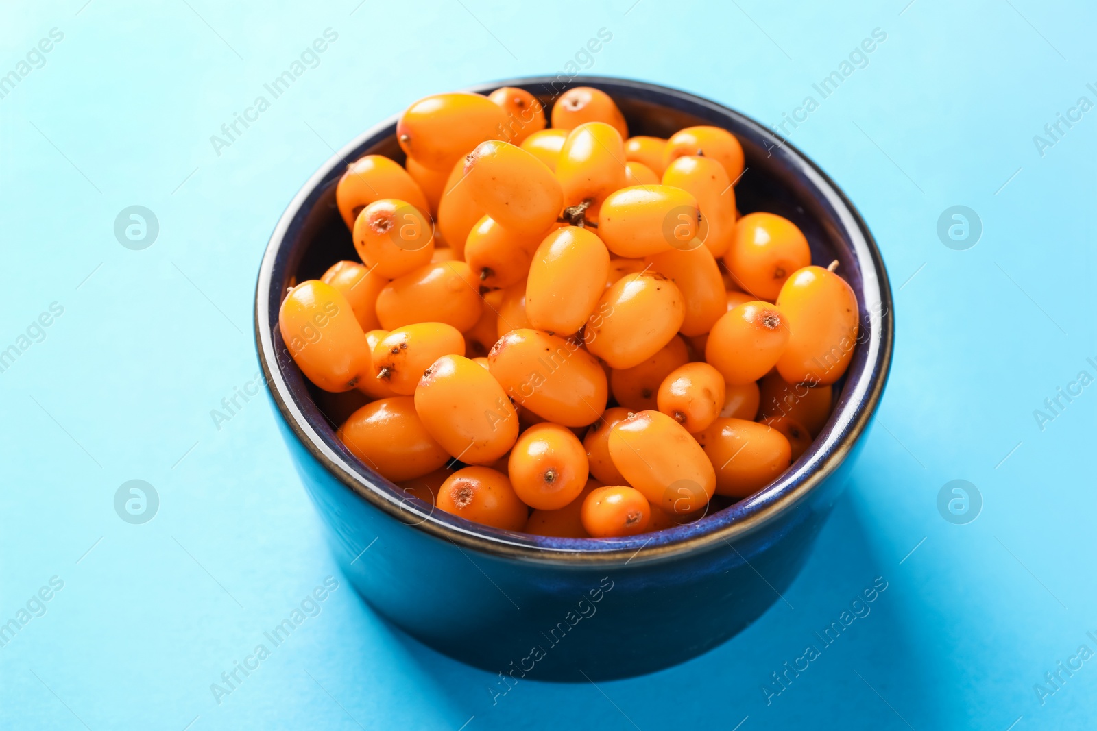 Photo of Ripe sea buckthorn berries in bowl on light blue background, closeup