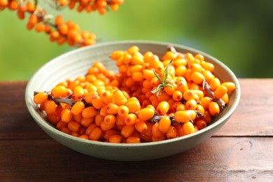 Photo of Ripe sea buckthorn berries and leaves in bowl on wooden table against blurred green background, closeup
