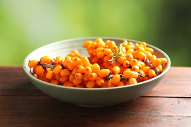 Photo of Ripe sea buckthorn berries and leaves in bowl on wooden table against blurred green background, closeup