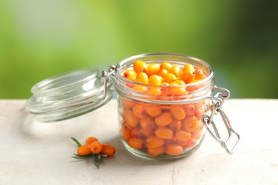 Photo of Ripe sea buckthorn berries in glass jar on light table against blurred green background, closeup