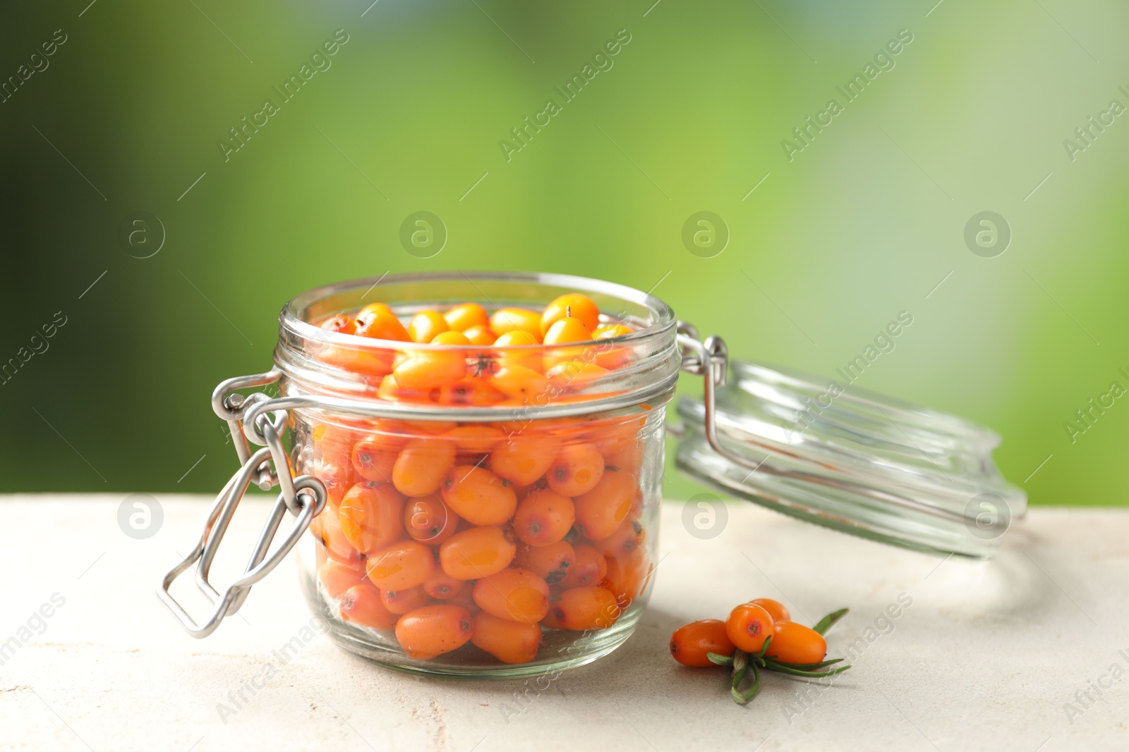 Photo of Ripe sea buckthorn berries in glass jar on light table against blurred green background, closeup