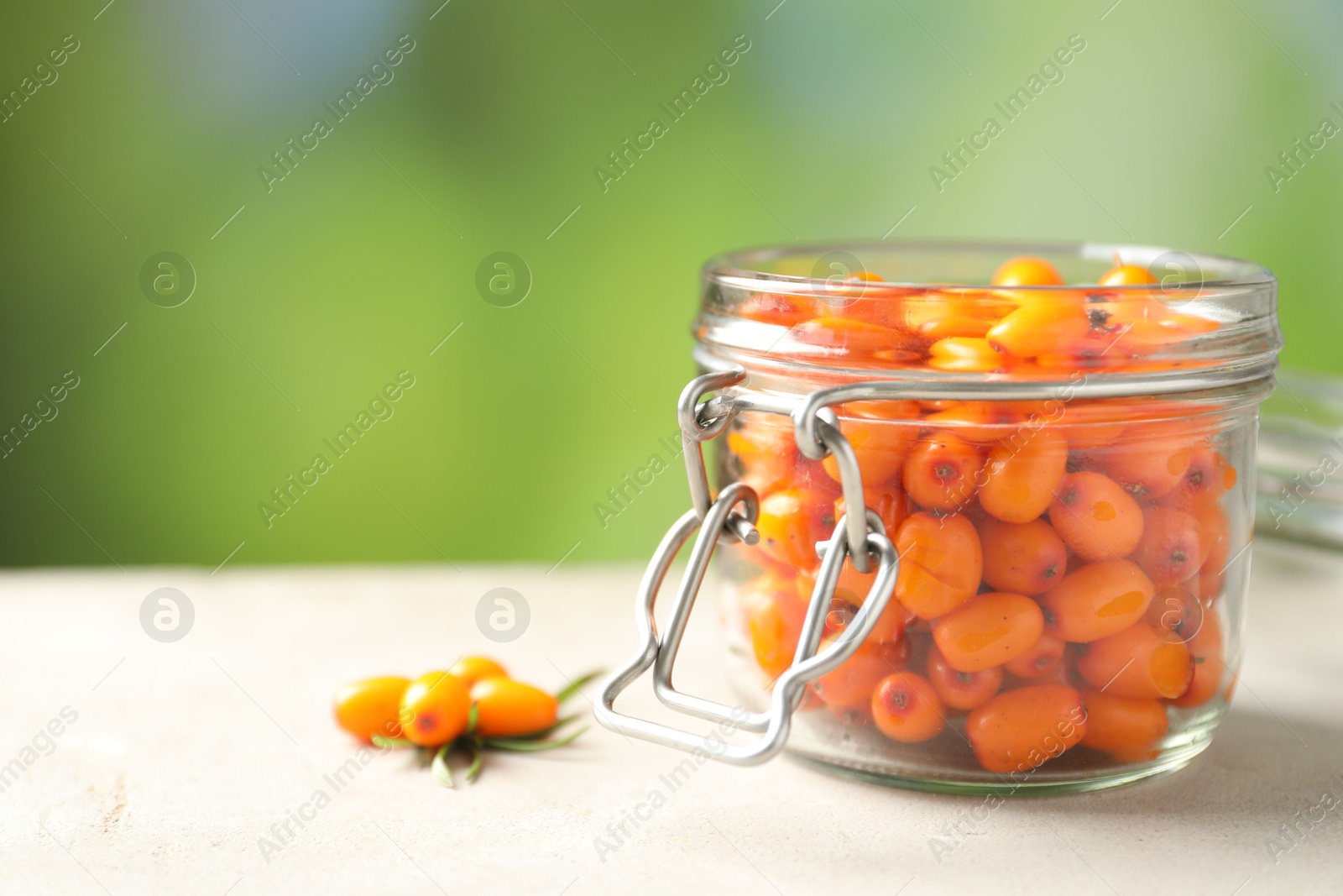 Photo of Ripe sea buckthorn berries in glass jar on light table against blurred green background, closeup. Space for text