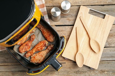 Photo of Air fryer with cooked chicken drumsticks and kitchen utensils on wooden table, flat lay