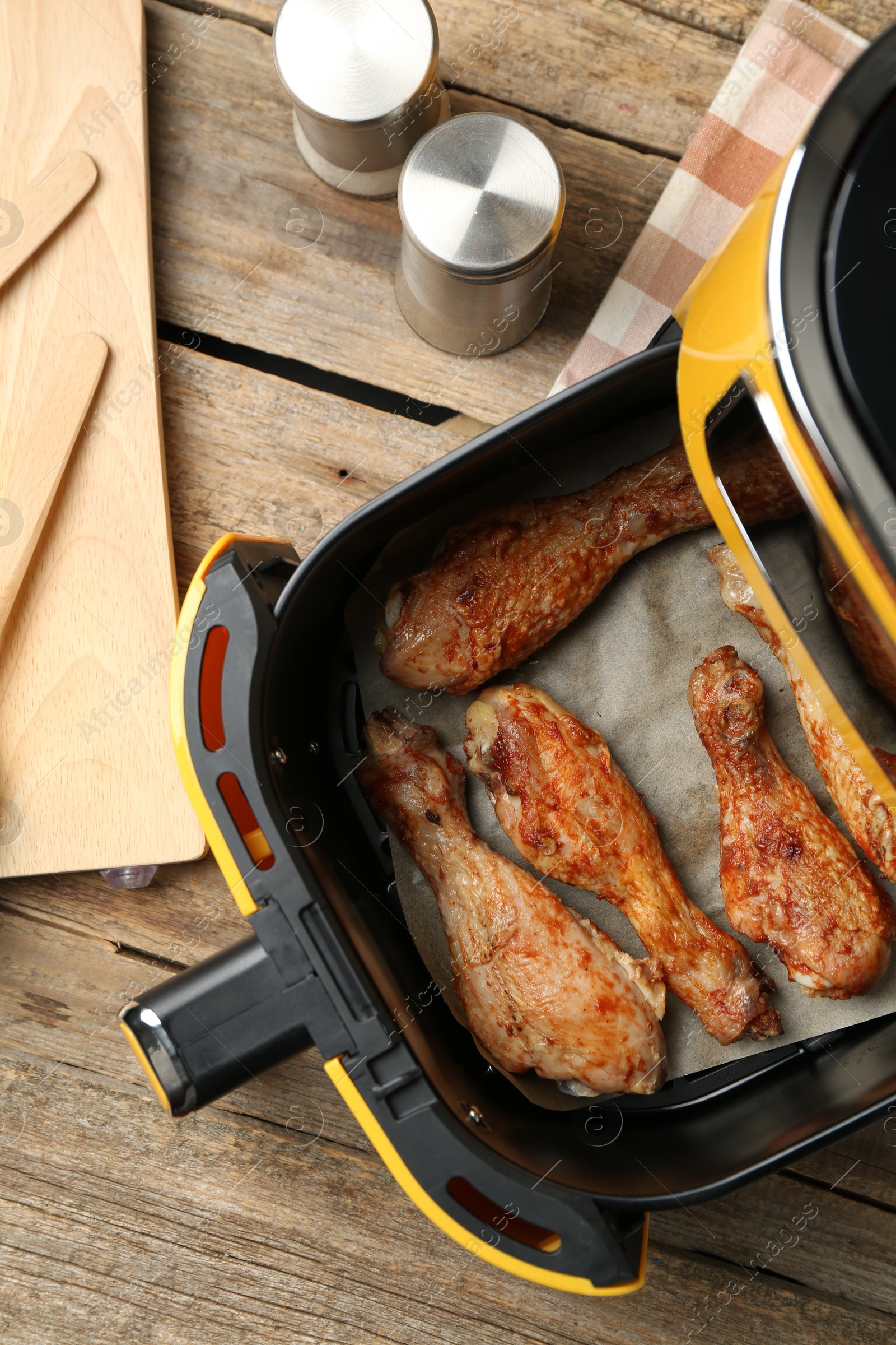 Photo of Air fryer with cooked chicken drumsticks and kitchen utensils on wooden table, flat lay