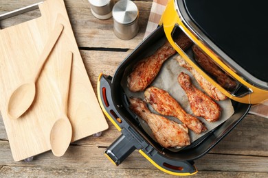Photo of Air fryer with cooked chicken drumsticks and kitchen utensils on wooden table, flat lay