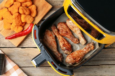 Photo of Air fryer with cooked chicken drumsticks and nuggets on wooden table, flat lay