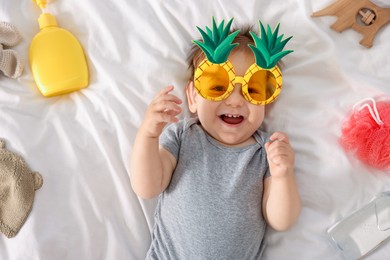 Photo of Cute baby with funny sunglasses and accessories on bed, top view