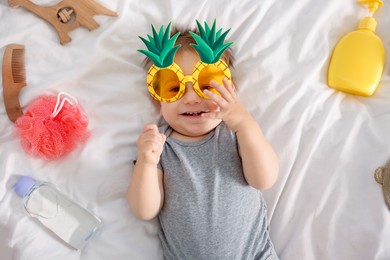 Photo of Cute baby with funny sunglasses and accessories on bed, top view