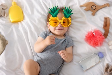 Photo of Cute baby with funny sunglasses and accessories on bed, top view