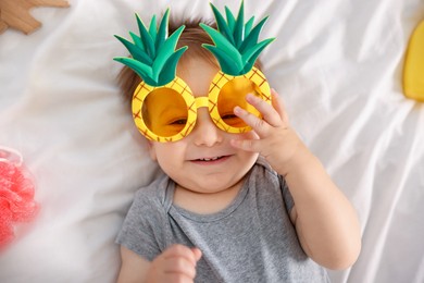 Photo of Cute baby with funny sunglasses and accessories on bed, top view