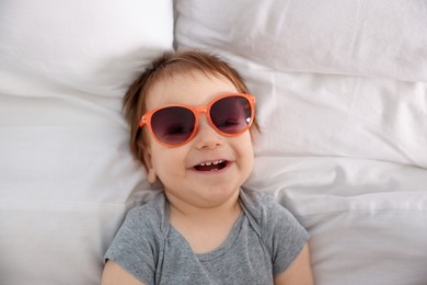 Photo of Funny baby with sunglasses lying on bed, top view