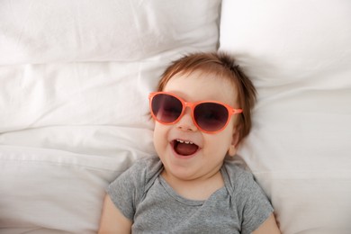 Photo of Funny baby with sunglasses lying on bed, top view