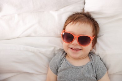Photo of Funny baby with sunglasses lying on bed, top view