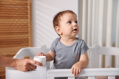 Photo of Mother with jar of cream and her baby in crib at home, closeup