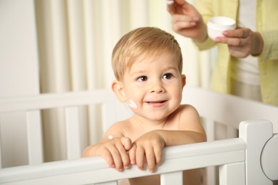 Photo of Cute baby with cream on face in crib and his mom at home