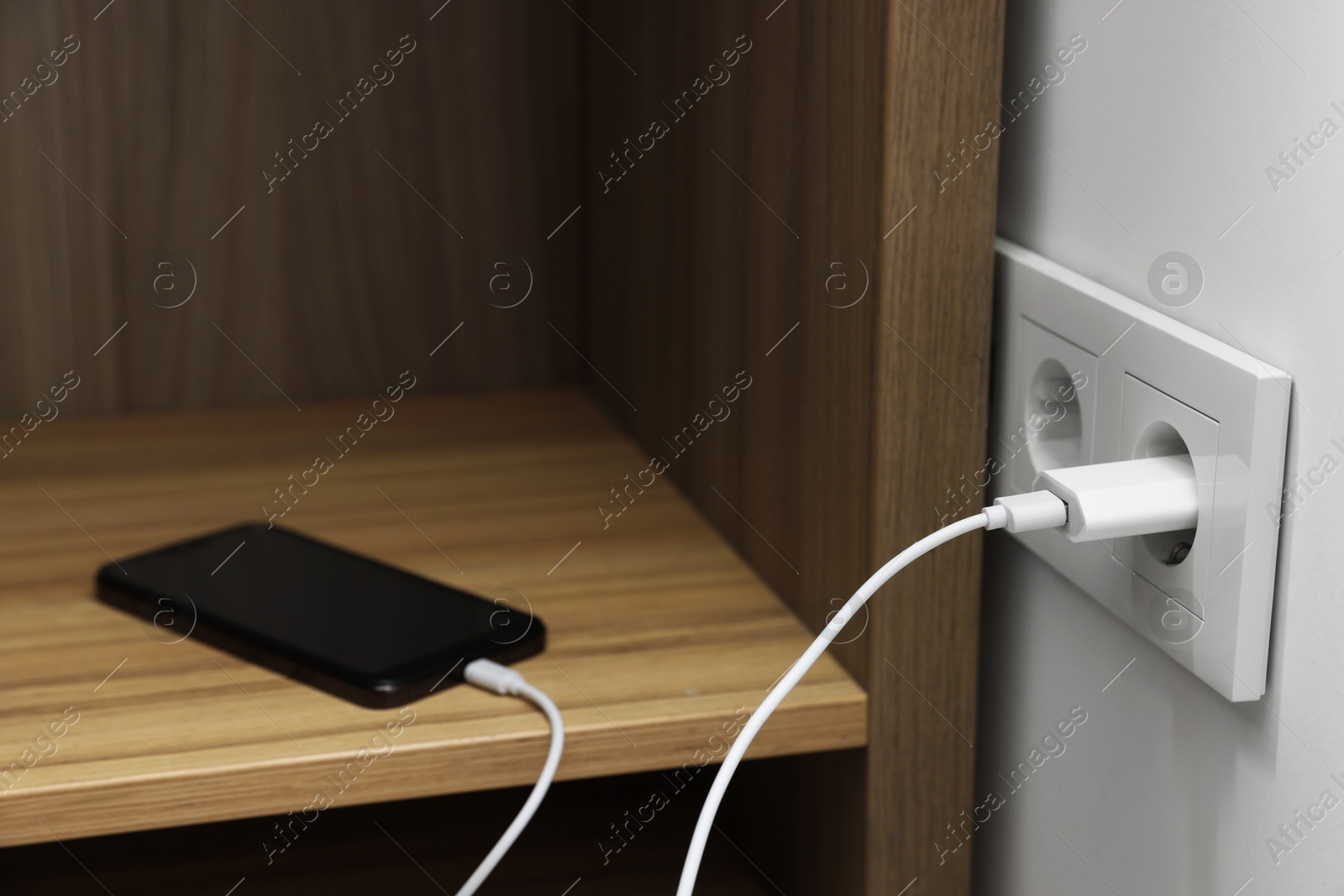 Photo of USB adapter with cable plugged into electric socket charging smartphone indoors, closeup