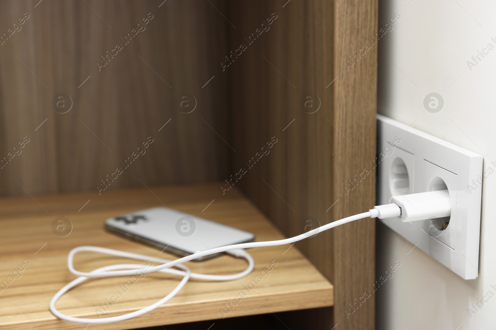 Photo of USB adapter with cable plugged into electric socket charging smartphone indoors, closeup