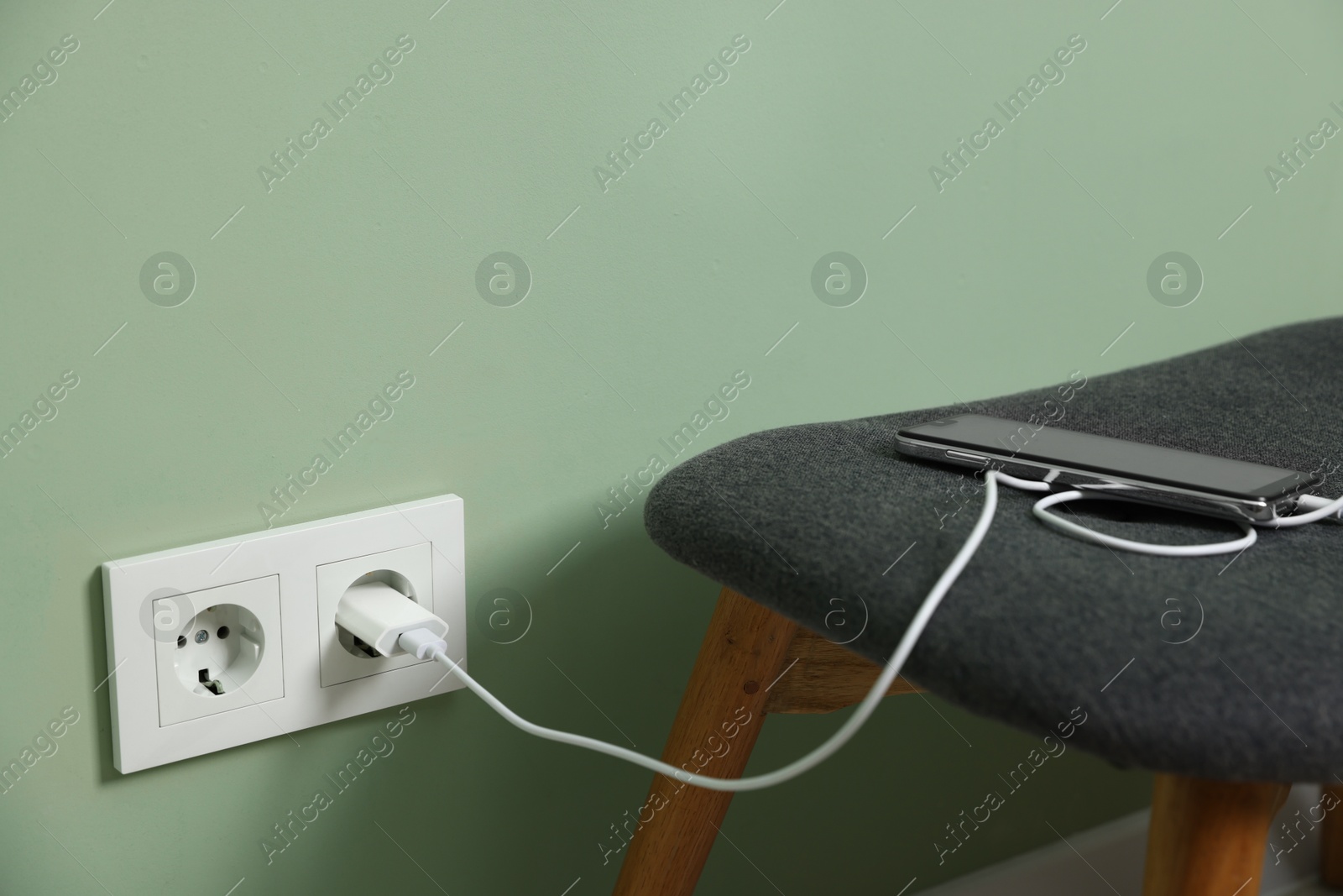 Photo of USB adapter with cable plugged into electric socket charging smartphone indoors