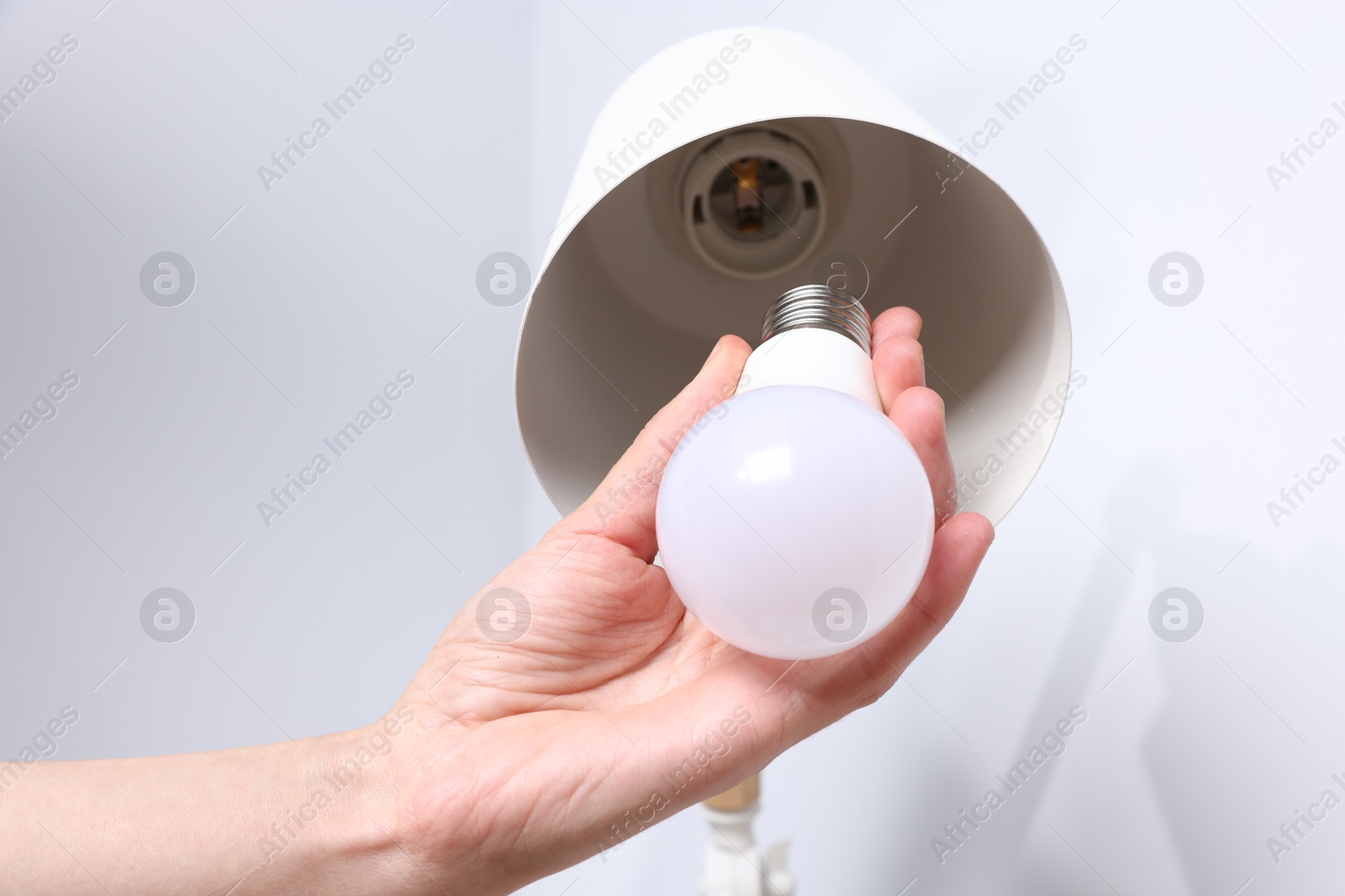 Photo of Woman changing light bulb in lamp near grey wall at home, closeup