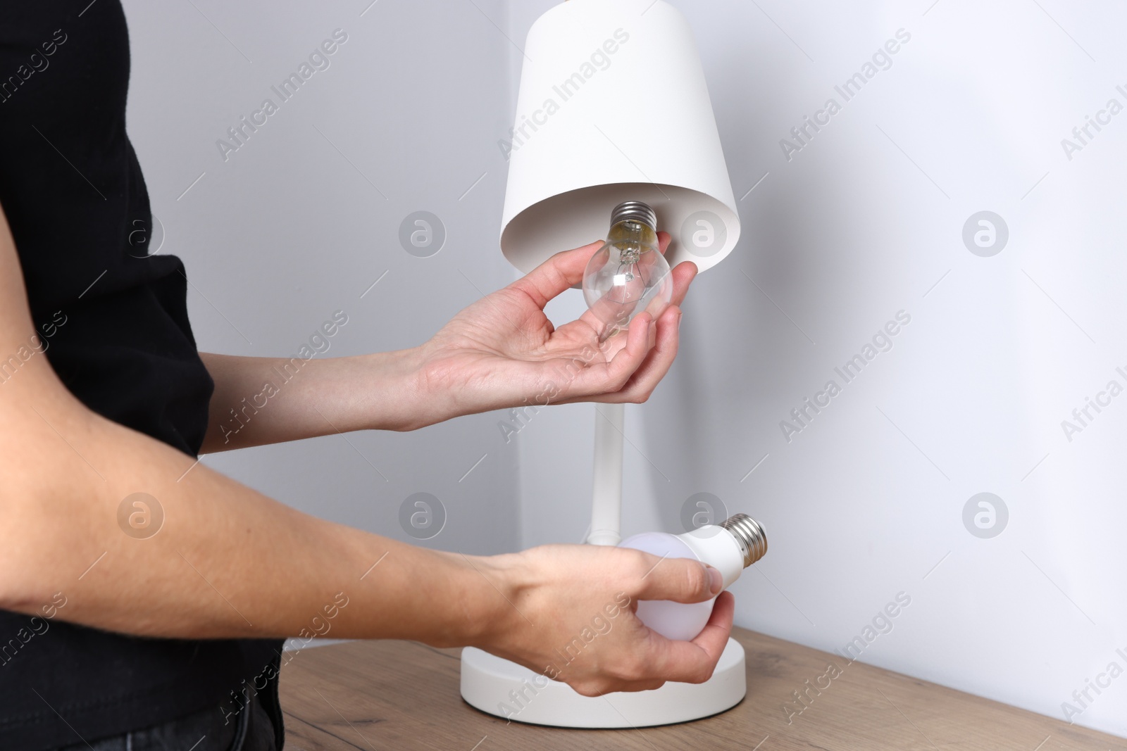 Photo of Woman changing light bulb in lamp near grey wall at wooden chest of drawers indoors, closeup