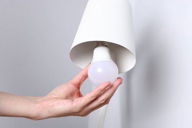 Photo of Woman changing light bulb in lamp near grey wall at home, closeup