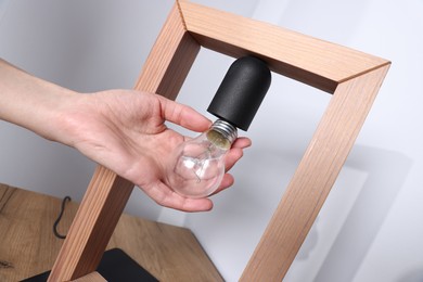 Photo of Woman changing light bulb in lamp near grey wall at wooden chest of drawers indoors, closeup