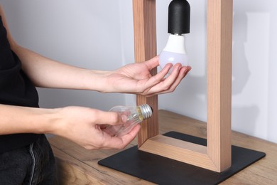 Photo of Woman changing light bulb in lamp near grey wall at wooden chest of drawers indoors, closeup