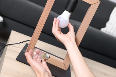 Photo of Woman changing light bulb in lamp at wooden table indoors, closeup