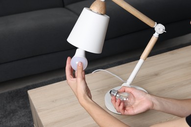 Photo of Woman changing light bulb in lamp at wooden table indoors, closeup