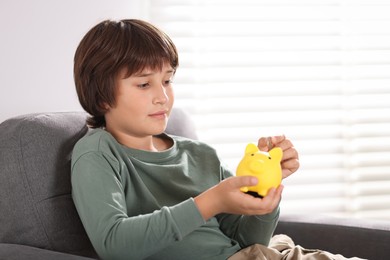Photo of Boy putting his pocket money into piggy bank in armchair at home. Space for text