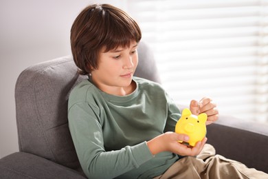 Photo of Boy putting his pocket money into piggy bank in armchair at home. Space for text