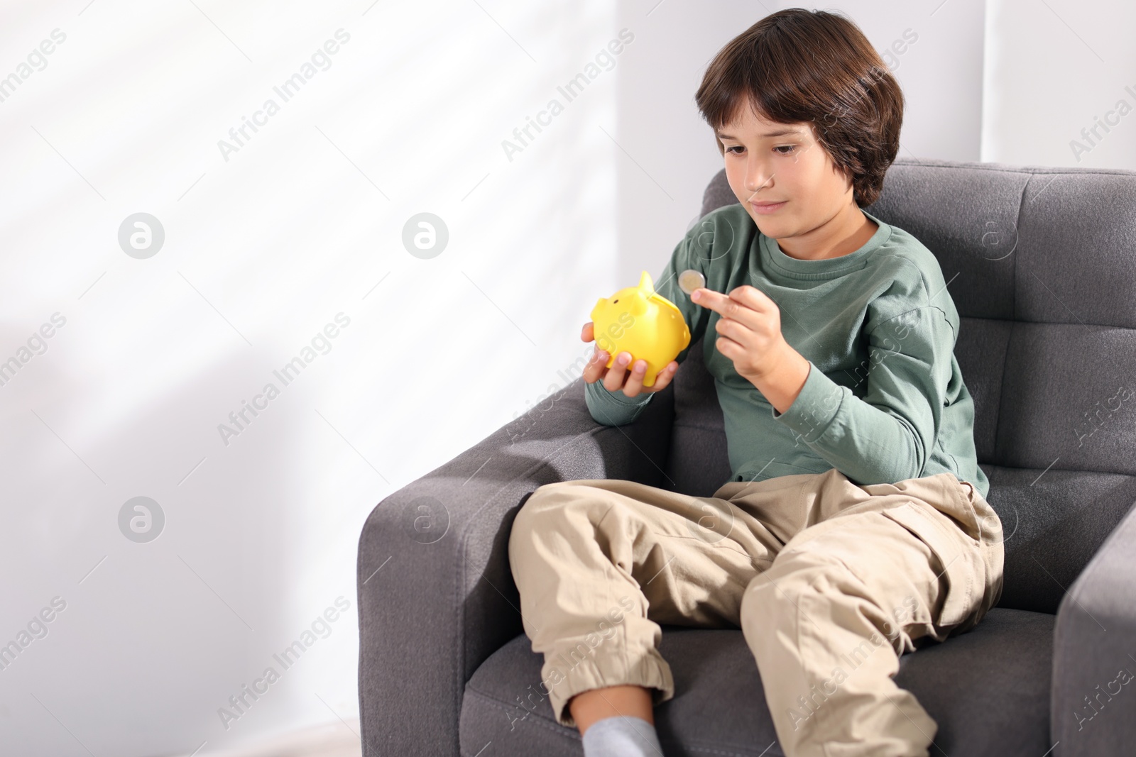 Photo of Boy putting his pocket money into piggy bank in armchair at home. Space for text