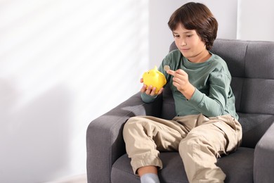 Photo of Boy putting his pocket money into piggy bank in armchair at home. Space for text