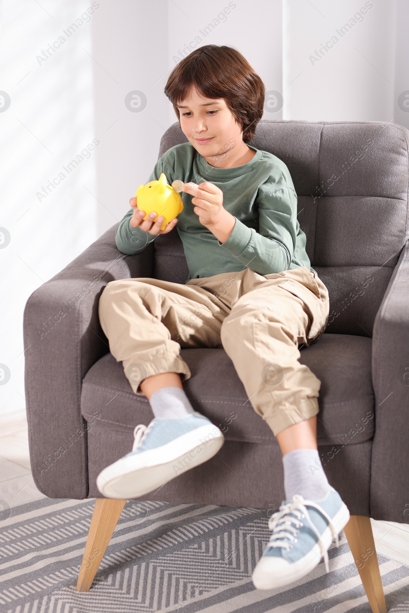 Photo of Boy putting his pocket money into piggy bank in armchair at home