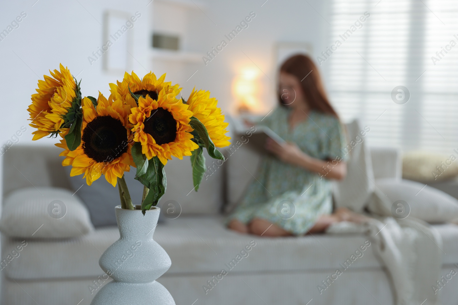 Photo of Girl reading book at home, focus on beautiful sunflowers in vase