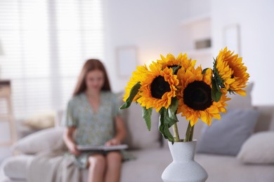 Photo of Girl reading book at home, focus on beautiful sunflowers in vase