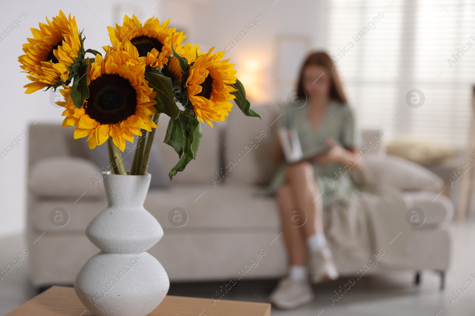 Photo of Girl reading book at home, focus on beautiful sunflowers in vase