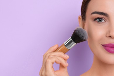 Image of Beautiful woman applying makeup with brush on light violet background, closeup. Space for text