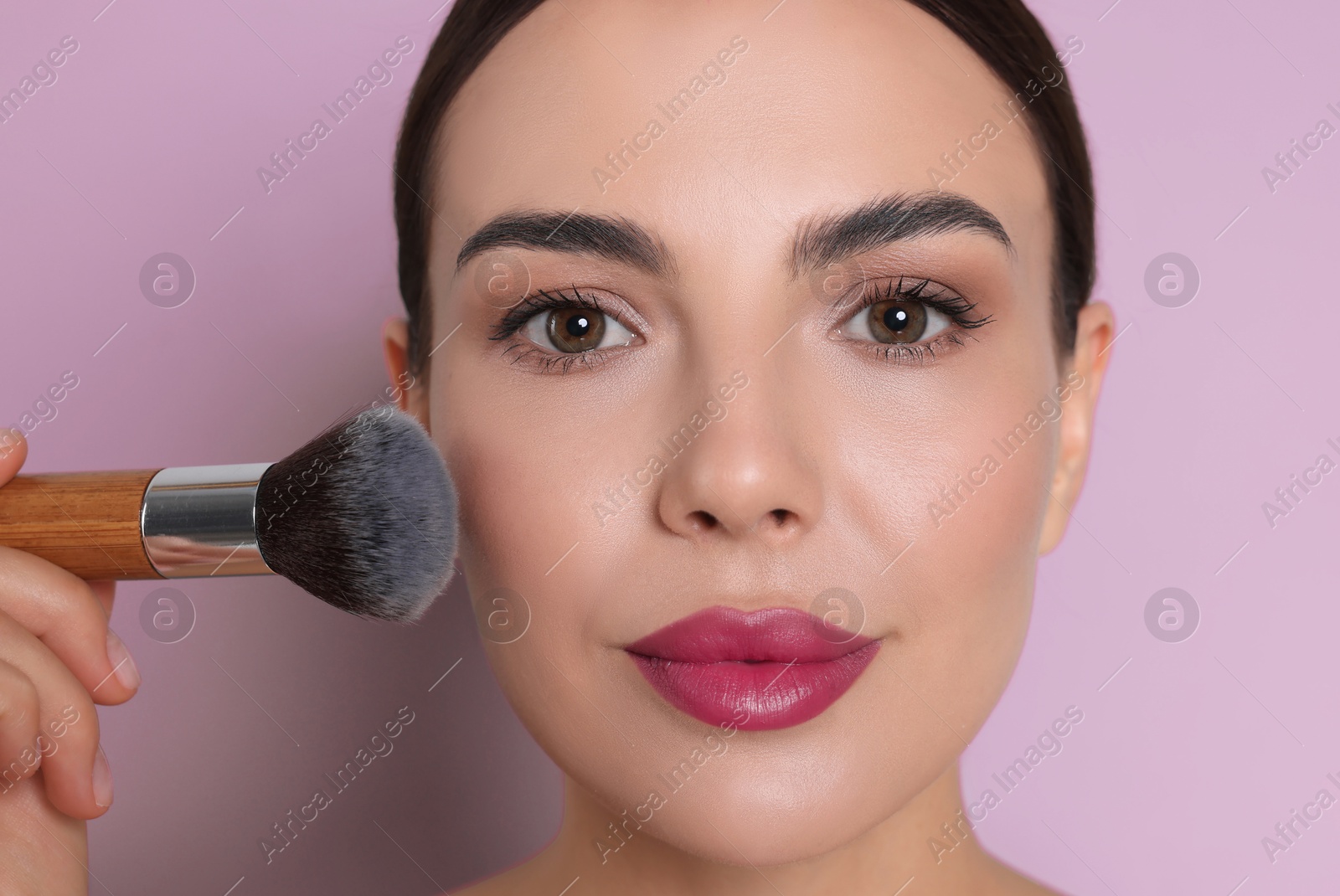 Image of Beautiful woman applying makeup with brush on lilac color background, closeup