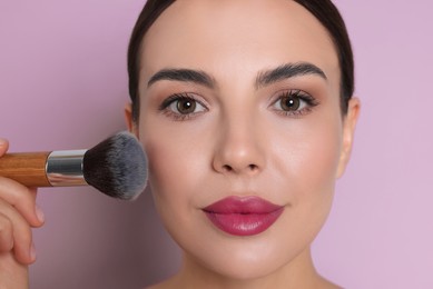 Image of Beautiful woman applying makeup with brush on lilac color background, closeup