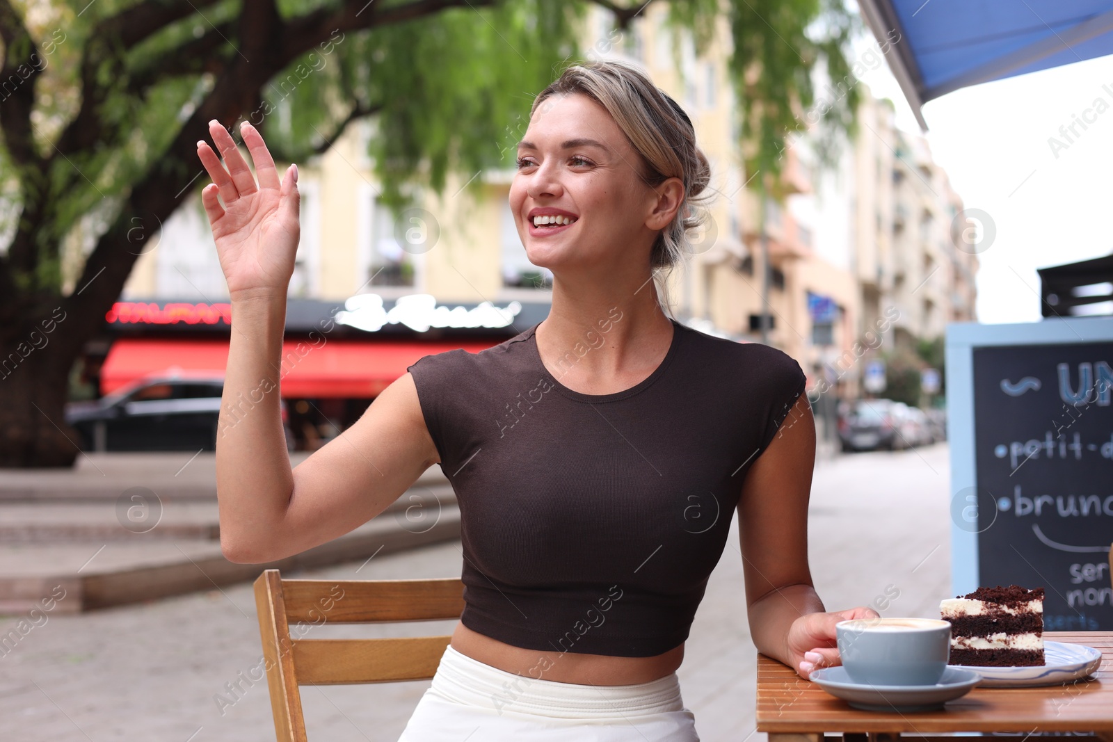 Photo of Beautiful young woman with cup of coffee and slice of cake at wooden table outdoors