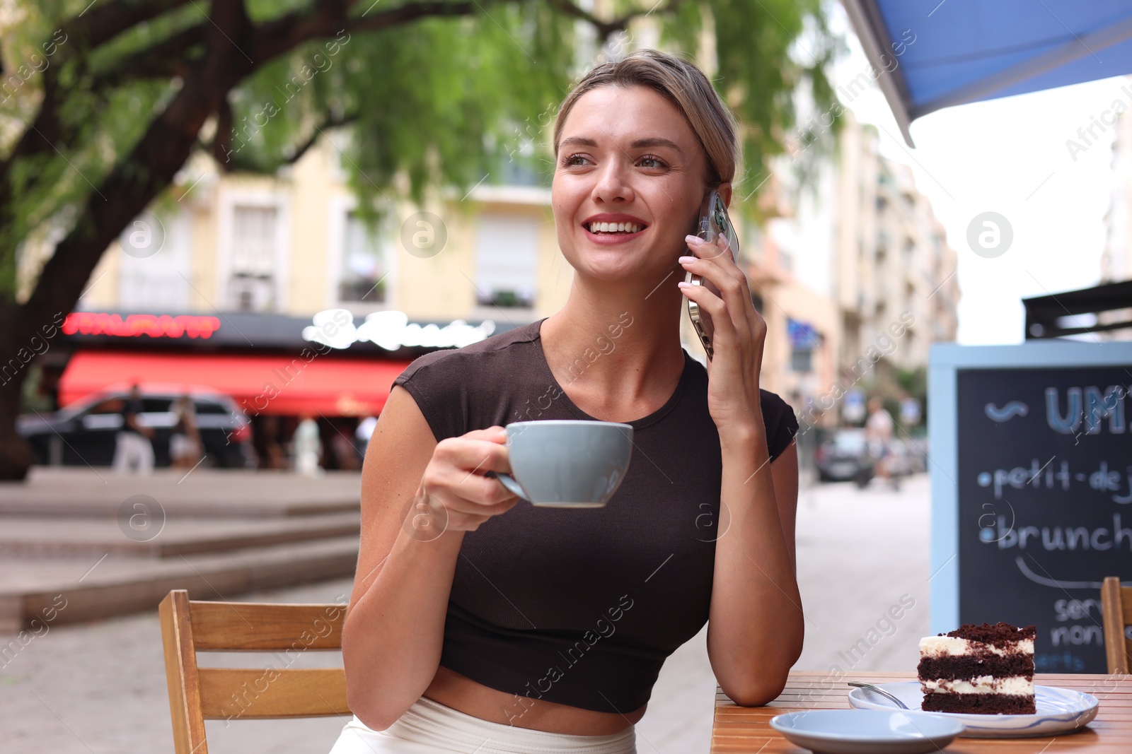 Photo of Beautiful young woman with cup of coffee and slice of cake talking on phone at wooden table outdoors