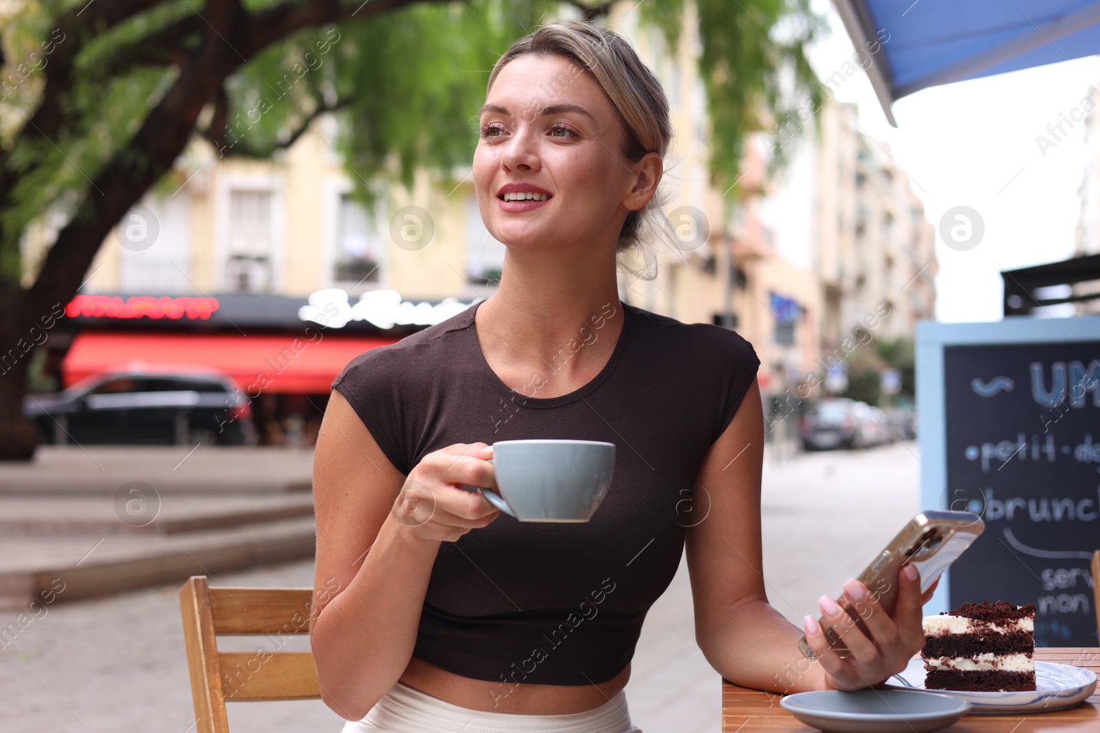 Photo of Beautiful young woman with cup of coffee, slice of cake and phone at wooden table outdoors