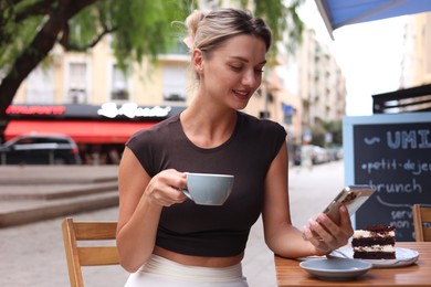 Photo of Beautiful young woman with cup of coffee, slice of cake and phone at wooden table outdoors