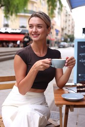 Photo of Beautiful young woman with cup of coffee and slice of cake at wooden table outdoors