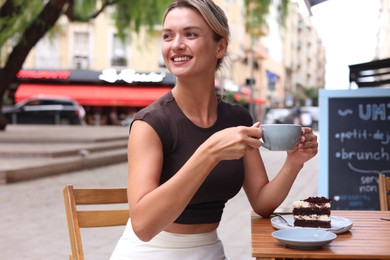 Photo of Beautiful young woman with cup of coffee and slice of cake at wooden table outdoors