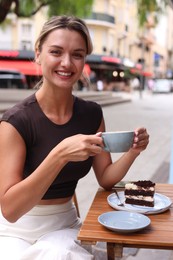 Photo of Beautiful young woman with cup of coffee and slice of cake at wooden table outdoors