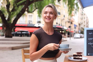 Photo of Beautiful young woman with cup of coffee and slice of cake at wooden table outdoors