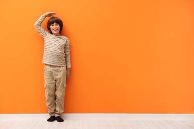 Photo of Cute boy measuring his height near orange wall indoors. Space for text